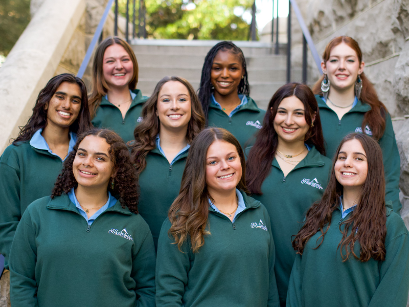 Nine smiling young women in green Hollabrook pullovers pose on stone steps outdoors.