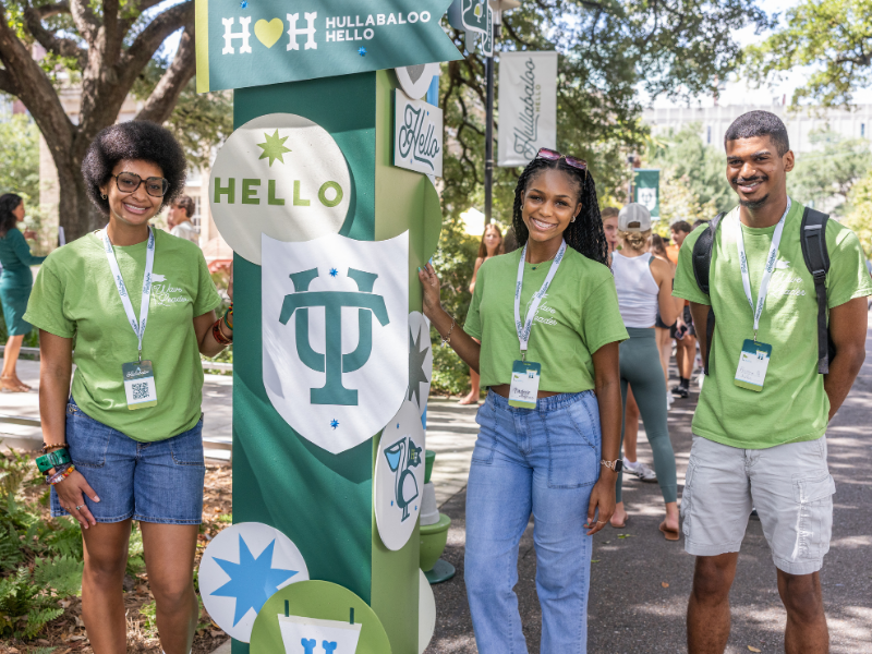 Three smiling Tulane Wave Leaders in green shirts next to a 'HULLABALOO HELLO' pillar on campus.