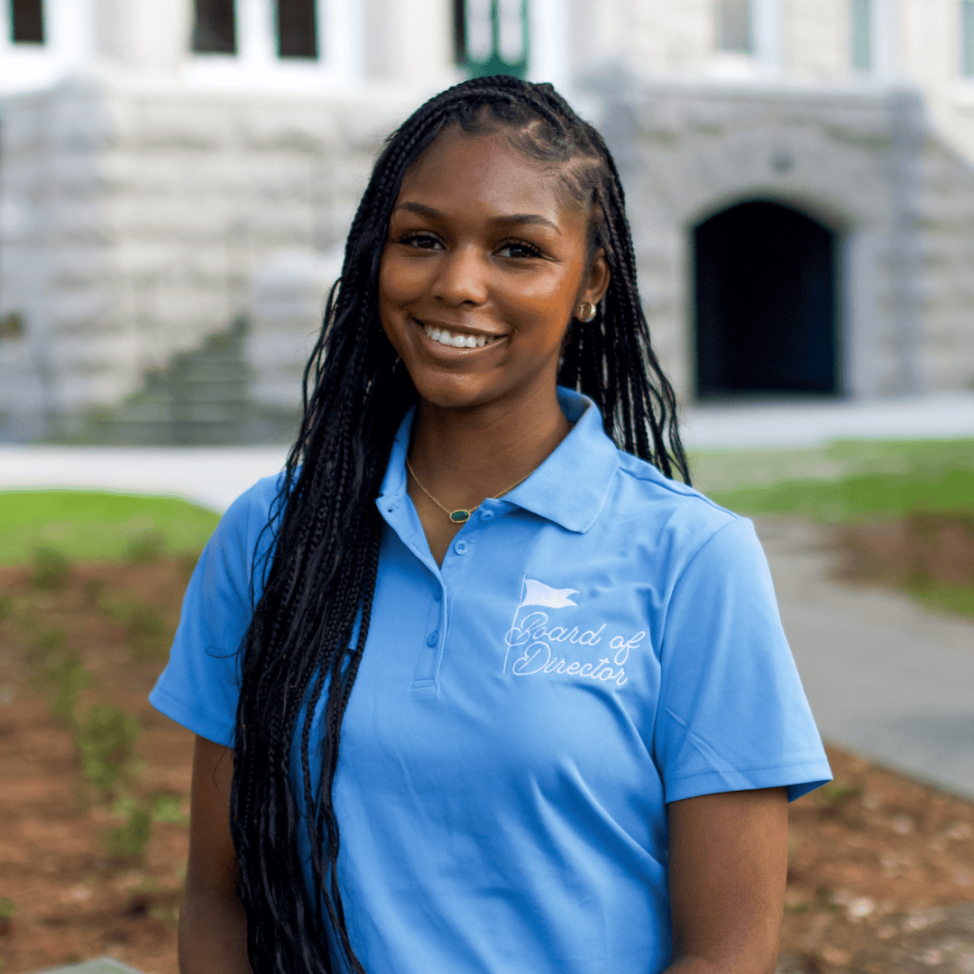 Young Black woman with braids smiling, wearing a blue polo shirt embroidered with "Board of Director" in front of a stone building.