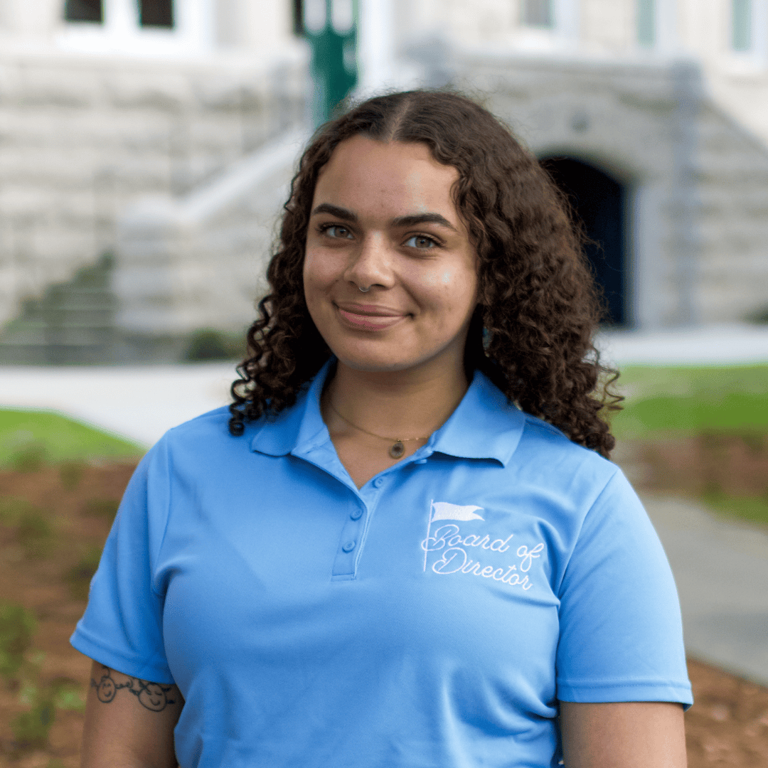 Smiling young woman with curly hair wearing a blue polo shirt with "Board of Director" logo.
