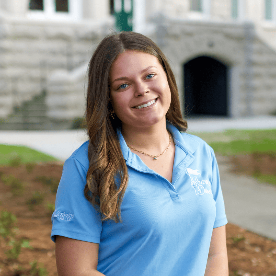 Smiling young woman in a light blue 'Boarding Dream' polo shirt with 'Callabalo's HELLO' on the sleeve, before a stone building.