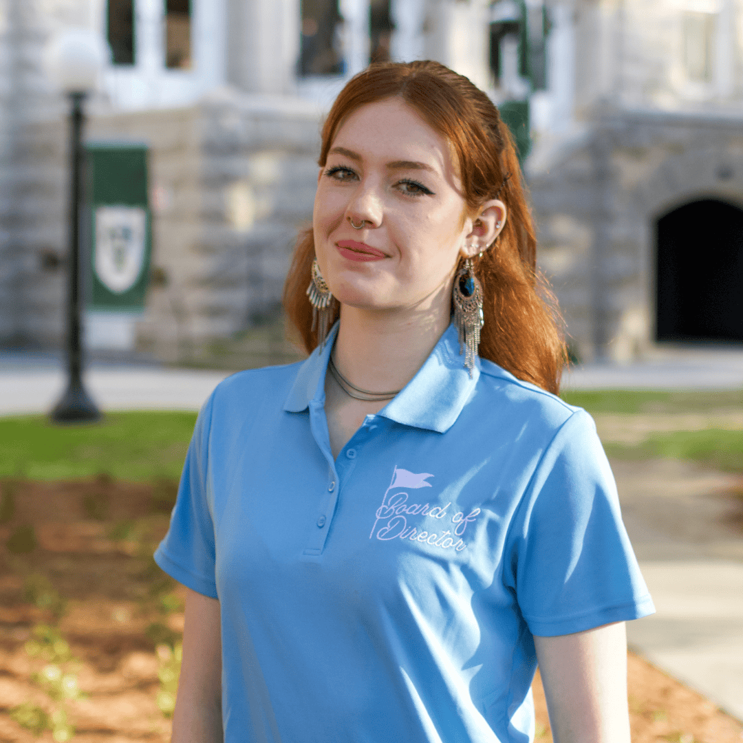 Young woman with a nose ring and long earrings wearing a light blue polo shirt with "Board of Director" logo, smiling outdoors.