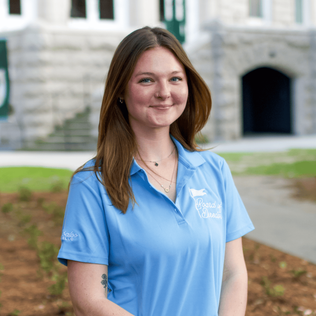 Young woman in a light blue polo shirt with "Board of Directors" and "Baloo ELLO" logos, smiling in front of a stone building.