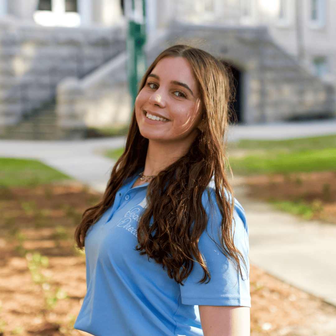 Young woman smiling in a light blue Coco Dreams polo shirt with a stone building and path in the background.