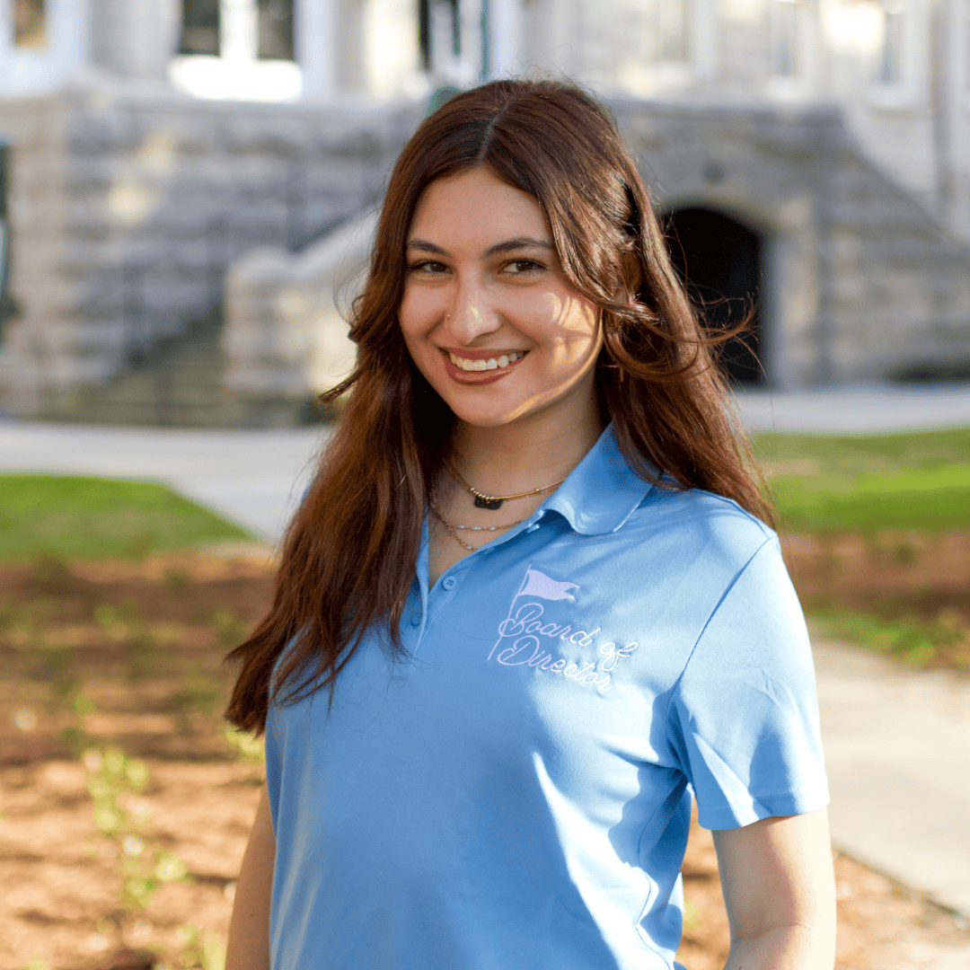 Smiling young woman in a light blue polo shirt with "Board of Director" text, standing outdoors near a stone building.