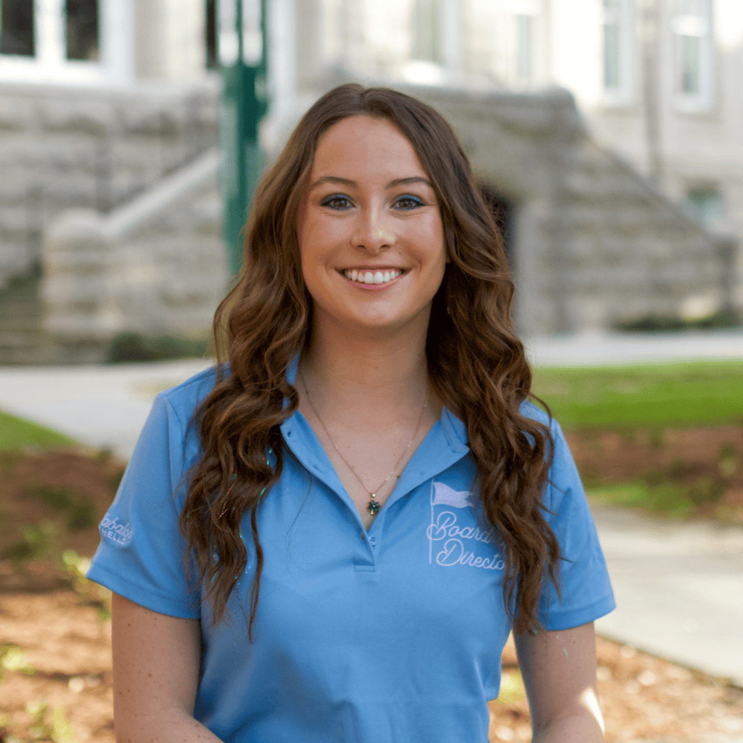Young woman smiling, wearing a light blue polo with a 'Board of Directors' logo and blue eyeliner, on a university campus.