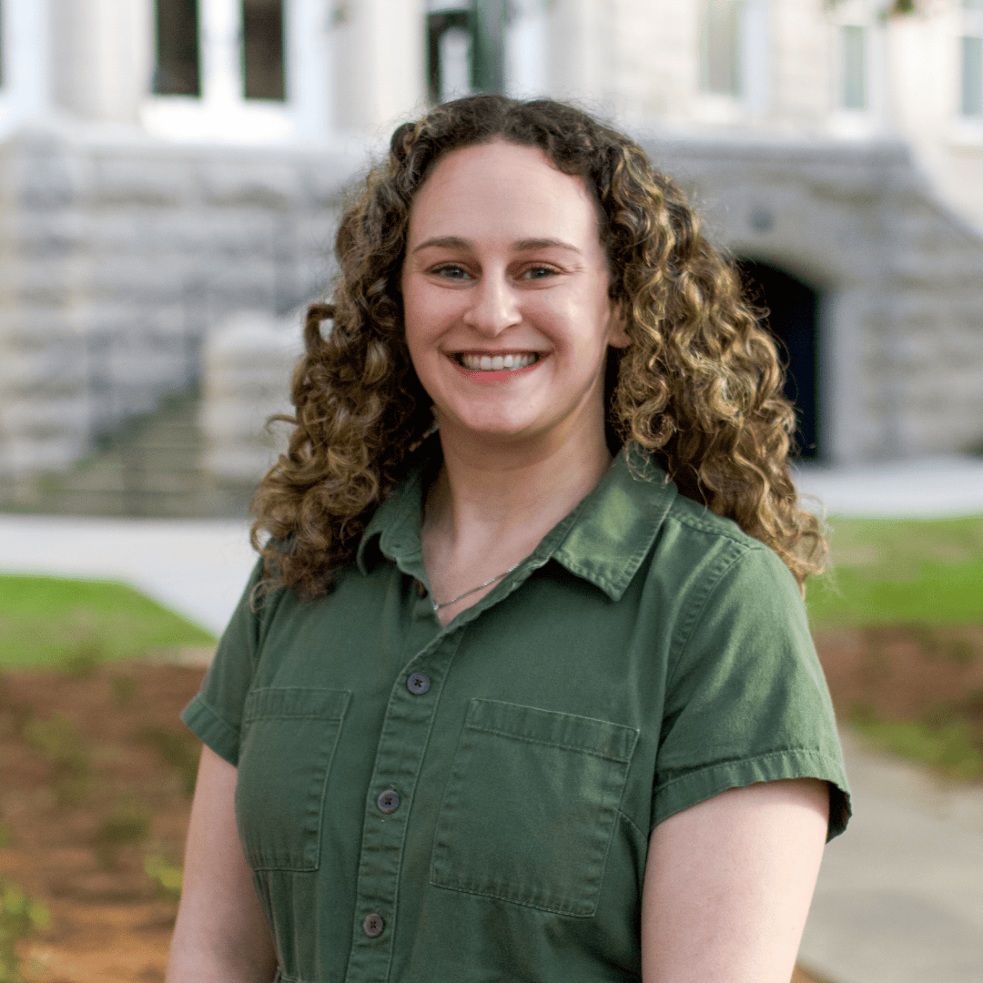 Smiling young woman with curly hair wearing a green shirt in front of a stone building.