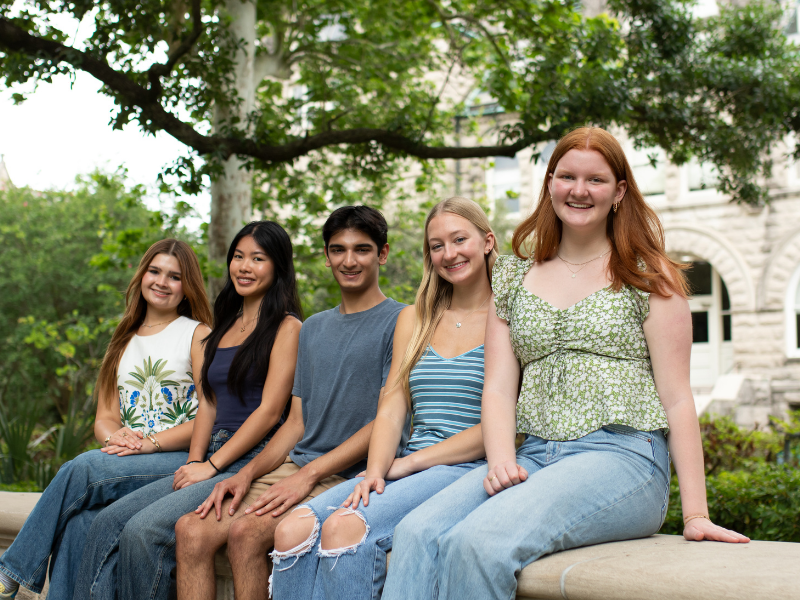 Five diverse smiling students sitting outdoors on a stone bench on a university campus.