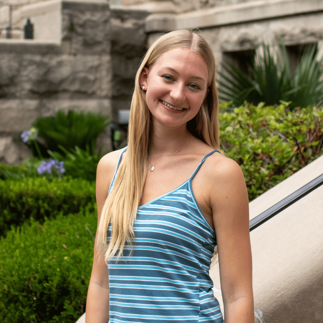 Smiling young woman with blonde hair and braces in a blue striped top outdoors.