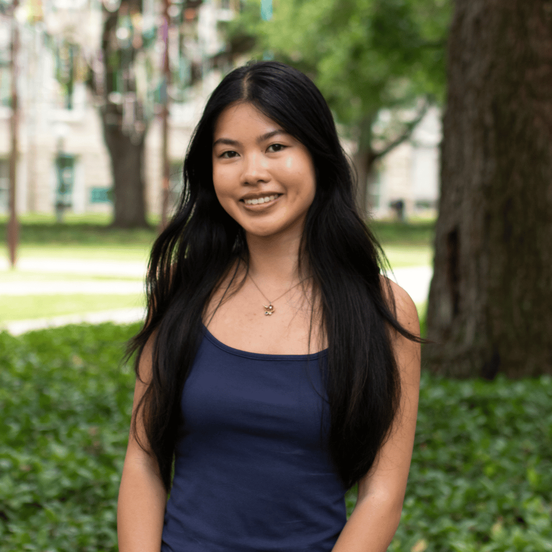 Smiling young Asian woman with long dark hair, wearing a navy tank top, stands outdoors with green foliage behind her.
